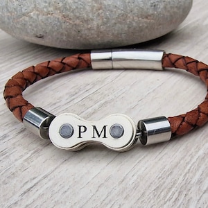 May include: Brown braided leather bracelet with a silver clasp and a decorative center piece. The center piece has the initials "PM" engraved on it. The bracelet is approximately 8 inches (20 cm) in length.