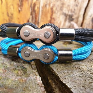 May include: Two bracelets, one black and one blue, featuring a bicycle chain link design. The bracelets have a braided rope band and silver-colored metal clasps. The chain links are stamped with the letters "KME".