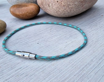 May include: A turquoise and grey rope bracelet with a silver magnetic clasp. The bracelet is circular and rests on a light-coloured wooden surface. The rope has a repeating pattern of turquoise and grey. The clasp is a shiny silver colour.