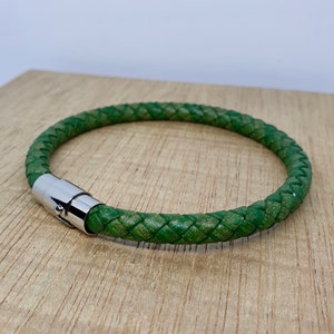May include: A green braided leather bracelet with a silver magnetic clasp. The bracelet is circular and appears to be approximately 8 inches in circumference. The leather has a textured appearance, and the clasp is shiny.