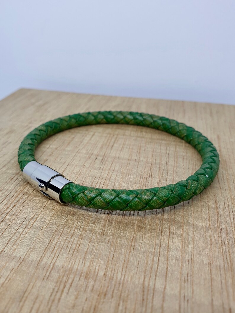 May include: A green braided leather bracelet with a silver magnetic clasp. The bracelet is circular and appears to be approximately 8 inches in circumference. The leather has a textured appearance, and the clasp is shiny.