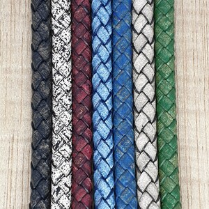 May include: A vertical close-up of seven braided leather cords in various colors. The colors include black, white with black speckles, burgundy, light blue, dark blue, gray, and green. The cords are arranged side by side.