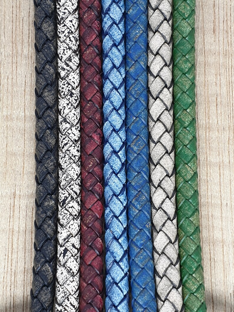 May include: A vertical close-up of seven braided leather cords in various colors. The colors include black, white with black speckles, burgundy, light blue, dark blue, gray, and green. The cords are arranged side by side.