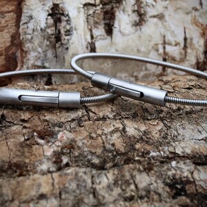 Pode incluir: Duas pulseiras de metal prateado e flex&iacute;vel com fecho de encaixe. As pulseiras s&atilde;o exibidas em uma superf&iacute;cie texturizada, marrom e bege. Os fechos s&atilde;o retangulares e t&ecirc;m um acabamento em metal escovado.