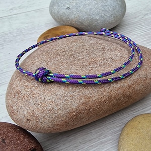 May include: A handmade bracelet with a purple, blue, and yellow cord. The bracelet is knotted in the centre and rests on a smooth, grey stone. The cord is approximately 0.5 cm in diameter.
