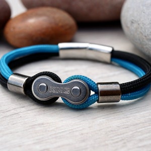 May include: A bracelet featuring a bicycle chain link design. The bracelet has a black and blue cord with silver-toned metal accents. The KMC logo is visible on the chain link. The bracelet is approximately 8 inches in circumference.