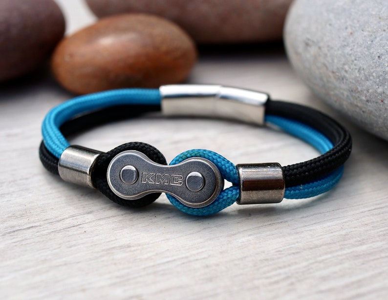 May include: A bracelet featuring a bicycle chain link design. The bracelet has a black and blue cord with silver-toned metal accents. The KMC logo is visible on the chain link. The bracelet is approximately 8 inches in circumference.