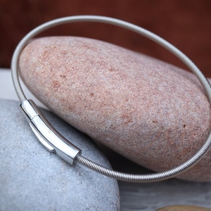 Pode incluir: Um colar prateado com um design flex&iacute;vel e enrolado. O colar apresenta um fecho retangular polido. A joia &eacute; exibida sobre uma pedra castanha clara, com uma pedra cinzenta em primeiro plano.