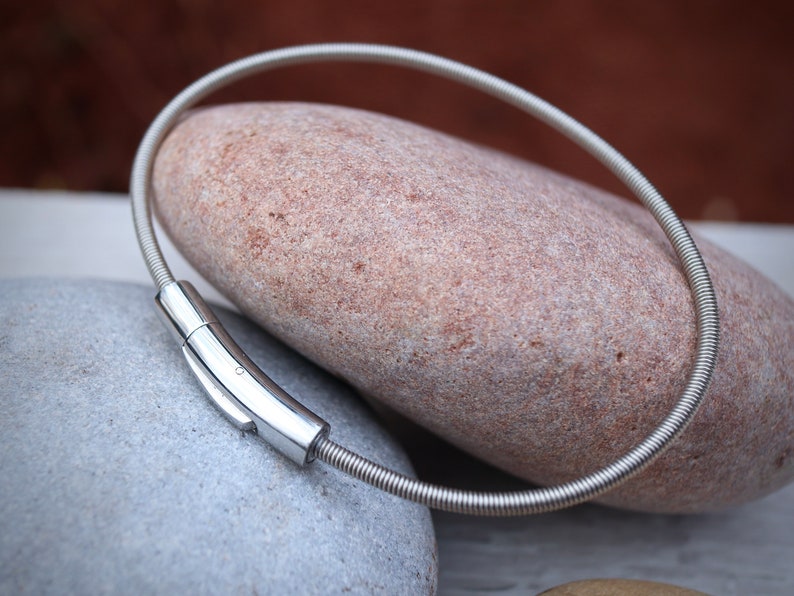 Pode incluir: Um colar prateado com um design flex&iacute;vel e enrolado. O colar apresenta um fecho retangular polido. A joia &eacute; exibida sobre uma pedra castanha clara, com uma pedra cinzenta em primeiro plano.