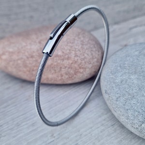 Pode incluir: Uma pulseira prateada com um design flex&iacute;vel e canelado. A pulseira apresenta um fecho retangular polido. A joia &eacute; exibida sobre uma superf&iacute;cie cinza claro com duas pedras no fundo.