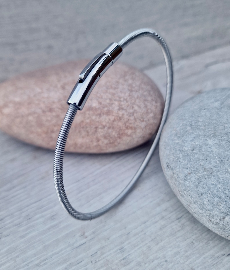 Pode incluir: Uma pulseira prateada com um design flex&iacute;vel e canelado. A pulseira apresenta um fecho retangular polido. A joia &eacute; exibida sobre uma superf&iacute;cie cinza claro com duas pedras no fundo.