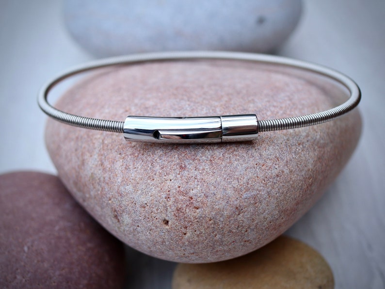 Pode incluir: Uma pulseira prateada com uma banda flex&iacute;vel e canelada e um fecho retangular polido. A pulseira est&aacute; sobre uma pedra lisa castanho-clara, com outras pedras no fundo.