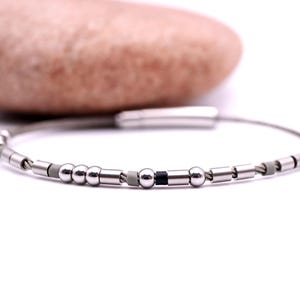 Può includere: Bracciale color argento con una serie di piccole perline cilindriche e sferiche. Il bracciale ha un design minimalista con una chiusura. Sullo sfondo c'è una pietra marrone chiaro.