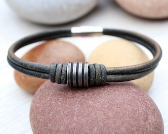 May include: A distressed leather bracelet with a silver-toned cylindrical clasp. The bracelet features two parallel leather cords in a muted green-brown hue. The clasp has vertical grooves. The bracelet is displayed on a natural stone surface.