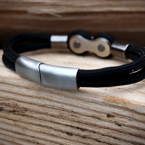 May include: A black braided rope bracelet with a silver-colored clasp and accents. The bracelet features a bicycle chain link design in the center. The bracelet is displayed on a wooden surface.