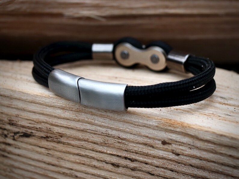 May include: A black braided rope bracelet with a silver-colored clasp and accents. The bracelet features a bicycle chain link design in the center. The bracelet is displayed on a wooden surface.