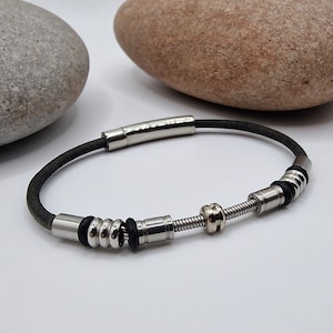 May include: A dark grey leather bracelet with silver metal detailing. The bracelet has a magnetic clasp and decorative metal beads. The design includes a coiled spring detail, adding a unique touch to the jewellery.