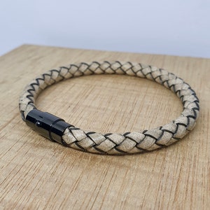 May include: Braided leather bracelet in a neutral beige color with black accents. The bracelet features a black magnetic clasp. The bracelet is displayed on a wooden surface.