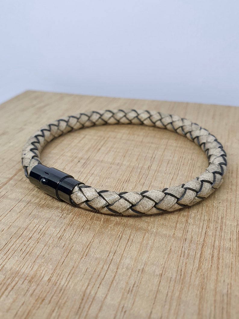 May include: Braided leather bracelet in a neutral beige color with black accents. The bracelet features a black magnetic clasp. The bracelet is displayed on a wooden surface.