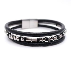 May include: A black leather bracelet with three strands. The bracelet has silver-coloured beads and a magnetic clasp. The design is simple and modern, suitable for everyday wear.