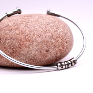 May include: A silver bangle bracelet featuring three small beads engraved with the words "sister", "mother", and "love". The bracelet is displayed on a light brown stone. The background is white.