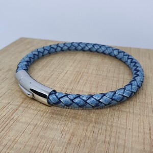 May include: A braided blue leather bracelet with a silver clasp. The bracelet is circular and appears to be made of woven leather strands. The clasp is a shiny, metallic silver. The bracelet is resting on a wooden surface.
