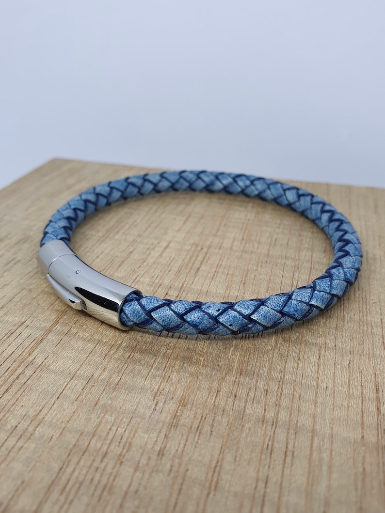 May include: A braided blue leather bracelet with a silver clasp. The bracelet is circular and appears to be made of woven leather strands. The clasp is a shiny, metallic silver. The bracelet is resting on a wooden surface.