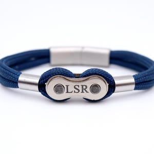 May include: A navy blue bracelet with a stainless steel clasp and a bicycle chain link design. The chain link is engraved with the letters "LSR". The bracelet is made of a woven rope material.