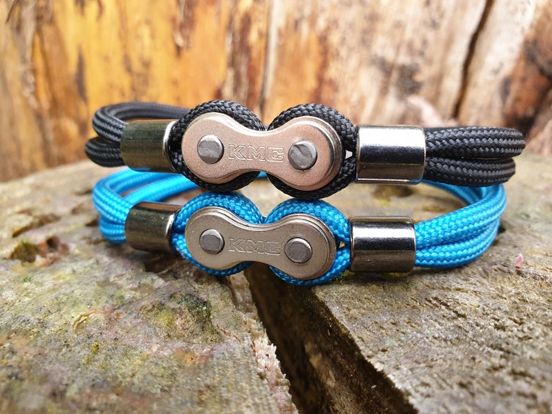 May include: Two bracelets, one black and one blue, featuring a bicycle chain link design. The bracelets have a braided rope band and silver-colored metal clasps. The chain links are stamped with the letters "KME".