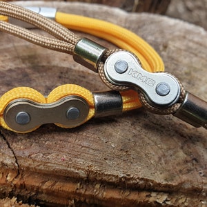 May include: Close-up of two bicycle chain bracelets. One bracelet features a tan cord and a silver chain link with the letters "KMC". The other bracelet has a yellow cord and a silver chain link. The background is a textured wooden surface.