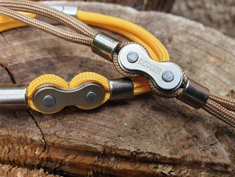 May include: Close-up of two bicycle chain bracelets. One bracelet features a tan cord and a silver chain link with the letters "KMC". The other bracelet has a yellow cord and a silver chain link. The background is a textured wooden surface.