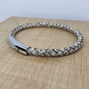 May include: Braided leather bracelet with a speckled black and white pattern. The bracelet features a polished silver clasp. The accessory is displayed on a light brown wooden surface.