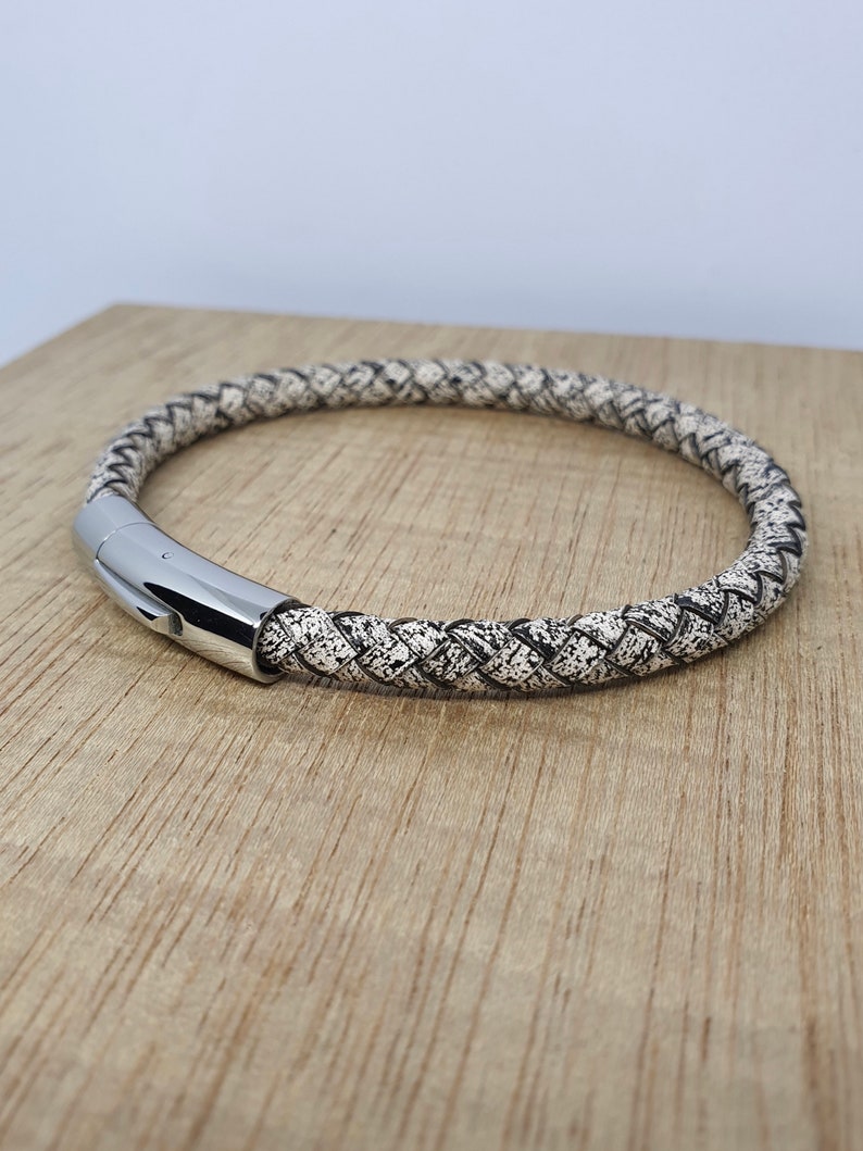May include: Braided leather bracelet with a speckled black and white pattern. The bracelet features a polished silver clasp. The accessory is displayed on a light brown wooden surface.