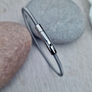 Pode incluir: Uma pulseira de metal prateado e flex&iacute;vel com um fecho. A pulseira &eacute; circular e parece ser feita de arame enrolado. O fecho tem formato retangular com uma pequena abertura. A pulseira &eacute; exibida em uma superf&iacute;cie de madeira clara.