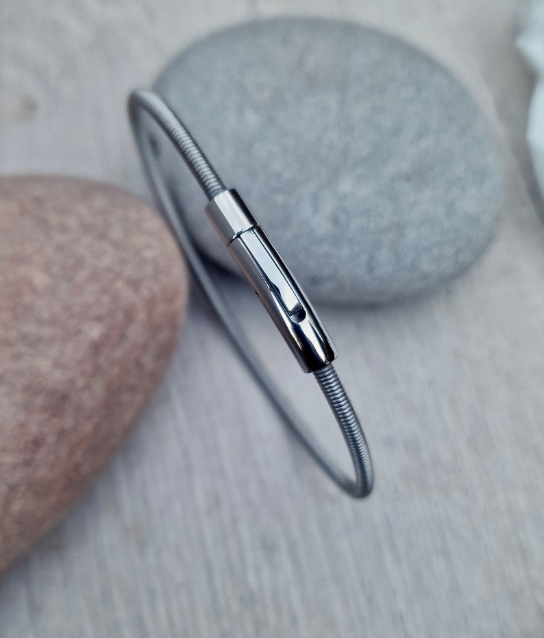 Pode incluir: Uma pulseira de metal prateado e flex&iacute;vel com um fecho. A pulseira &eacute; circular e parece ser feita de arame enrolado. O fecho tem formato retangular com uma pequena abertura. A pulseira &eacute; exibida em uma superf&iacute;cie de madeira clara.