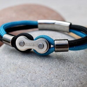 May include: A bicycle chain bracelet with a KMC logo. The bracelet features a silver-colored chain link and magnetic clasp, with blue and black braided cord. The bracelet is a stylish accessory for cyclists and those who appreciate industrial design.