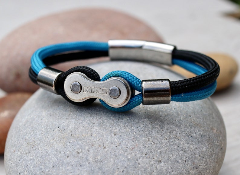 May include: A bicycle chain bracelet with a KMC logo. The bracelet features a silver-colored chain link and magnetic clasp, with blue and black braided cord. The bracelet is a stylish accessory for cyclists and those who appreciate industrial design.