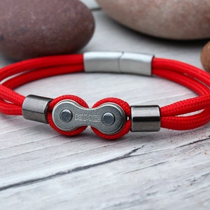 May include: A red bracelet with a bicycle chain link clasp. The bracelet features a bright red cord and a silver magnetic clasp. The bicycle chain link is silver with the letters "KMC" engraved on it. The bracelet is on a light wood surface.