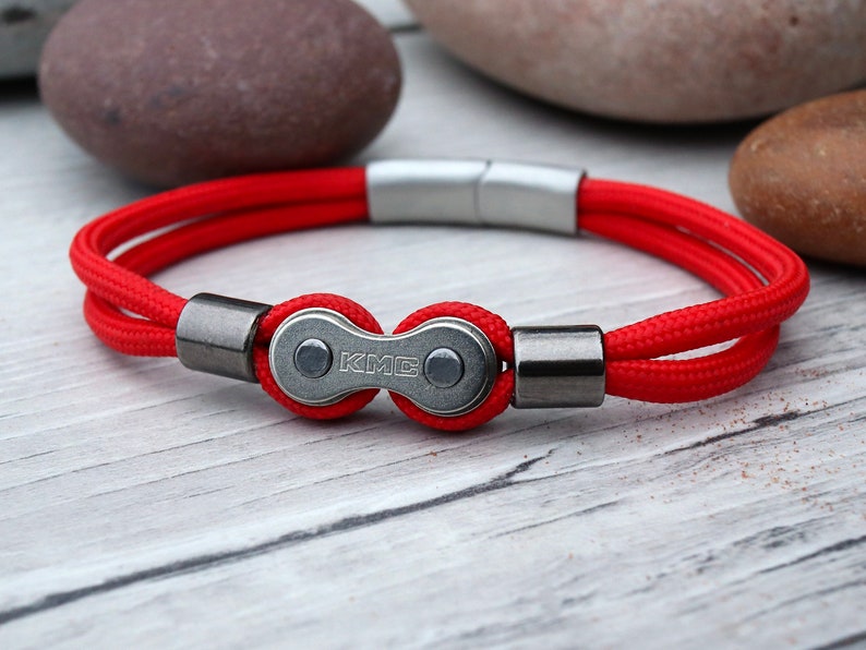 May include: A red bracelet with a bicycle chain link clasp. The bracelet features a bright red cord and a silver magnetic clasp. The bicycle chain link is silver with the letters "KMC" engraved on it. The bracelet is on a light wood surface.