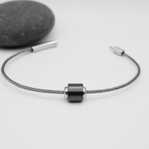 May include: A minimalist bracelet featuring a dark gray cable with a cylindrical, dark gray bead at the center. The bracelet has silver-toned end caps. A gray stone sits in the background on a white surface.