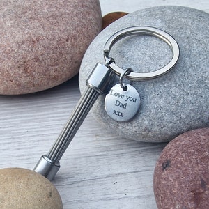 May include: A silver-toned keychain with a spring-like design and a round charm. The charm is engraved with the words "Love you Dad" and "xxx". The keychain is shown against a background of smooth, gray and brown stones.