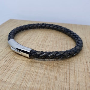 May include: A braided black leather bracelet with a silver-colored magnetic clasp. The bracelet is displayed on a light brown wooden surface. The leather has a textured appearance.