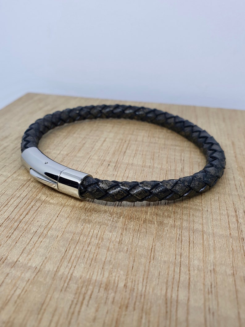 May include: A braided black leather bracelet with a silver-colored magnetic clasp. The bracelet is displayed on a light brown wooden surface. The leather has a textured appearance.