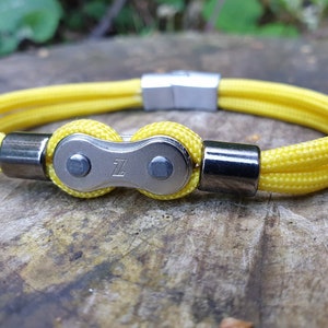 May include: Yellow rope bracelet with a silver-colored bicycle chain link design. The bracelet has a magnetic clasp and metal accents. The bicycle chain link has the letter "Z" engraved on it.