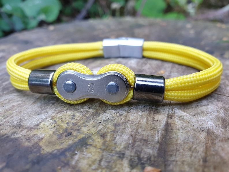 May include: Yellow rope bracelet with a silver-colored bicycle chain link design. The bracelet has a magnetic clasp and metal accents. The bicycle chain link has the letter "Z" engraved on it.
