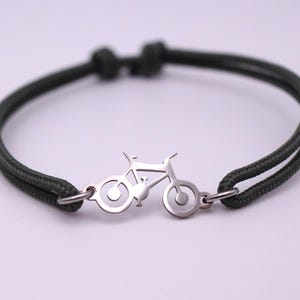 May include: A dark green bracelet with a silver bicycle charm. The bracelet features a double-strand cord design and a small, detailed bicycle silhouette in the center. The clasp is a simple loop and ring design.