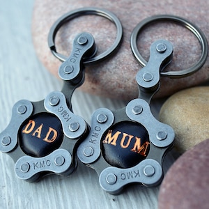 May include: Two keychains made from bicycle chain links. One keychain has the word "DAD" in gold lettering on a black background. The other keychain has the word "MUM" in gold lettering on a black background. Both keychains have silver key rings.