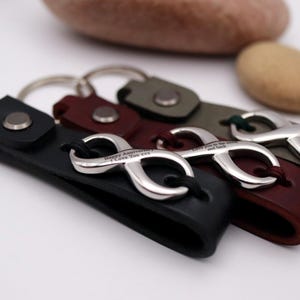 May include: Leather keyrings in black, burgundy, and olive green, each with a silver infinity symbol. The infinity symbols have engraved text. The keyrings are displayed on a white surface with decorative stones in the background.