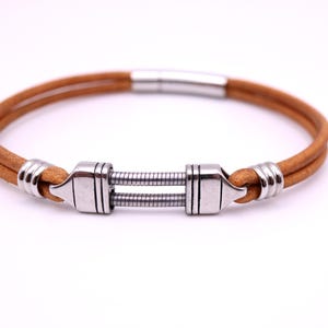 Peut inclure: Un bracelet en cordon de cuir beige composé de deux brins parallèles. Le bracelet présente des éléments en métal argenté, notamment deux perles cylindriques striées et une pièce centrale avec deux ressorts métalliques fins et enroulés.