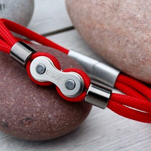 May include: Red rope bracelet featuring a silver-colored bicycle chain link with the letters "KMC". The bracelet has silver-colored metal end caps and a magnetic clasp. The bracelet is displayed on a wooden surface with rocks in the background.